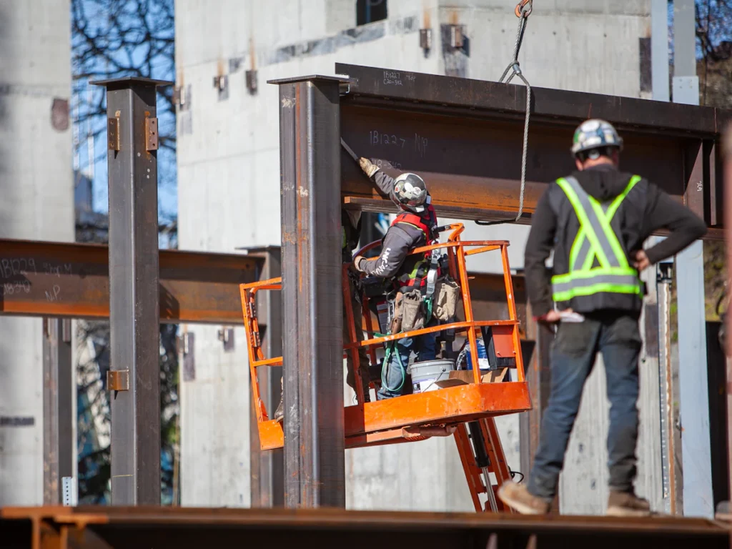 Ironworkers in safety vests and hard hats setting steel from a lift, the level-by-level installation used for fire escape replacement in Westchester